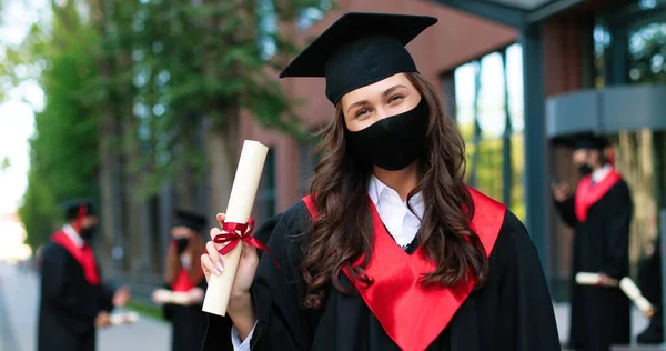 Portrait of caucasian female student wearing black hat and protective mask posing to the camera and showing her diploma after graduated from university during the covid 19 pandemic. Education concept — ストック写真