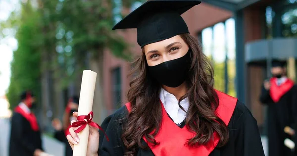 Portrait of caucasian female student wearing black hat and protective mask posing to the camera and showing her diploma after graduated from university during the covid 19 pandemic. Education concept — ストック写真