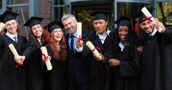 Gesellschaft fröhlicher, multiethnischer Studenten feiert ihren Abschluss, während sie in der Nähe der Hochschule stehen und mit Diplomen in der Hand jubeln. Bildungskonzept — Stockfoto