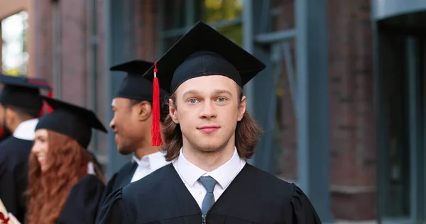 Graduation. Portrait view of the caucasian happy boy looking to the camera with wide smile while standing with his group mates and listening final speech from his teacher. Education concept