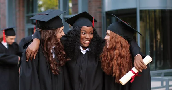 Glückliche Gruppe reifer Studenten am Abschlusstag, die sich umarmen. Drei beste Freundinnen in akademischen Kleidern und Mützen umarmen sich vor der Kamera — Stockfoto