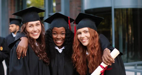 Щаслива група дорослих студентів у день випуску, що охоплює один одного. Три найкращих подруги-дівчата в академічних сукнях і шапках обіймаються перед камерою — стокове фото