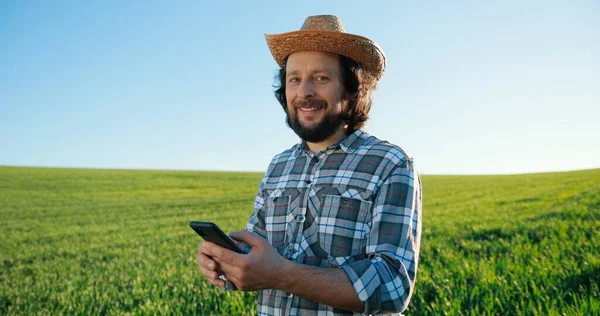 Waist up portrait view of the optimistic caucasian farmer scientist in hat using smartphone mobile technology application while working at the field and smiling. Agriculture concept — Stock Photo, Image