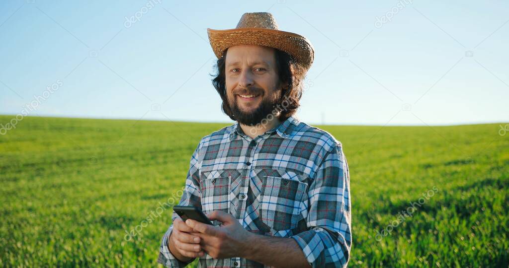 Waist up portrait view of the optimistic caucasian farmer scientist in hat using smartphone mobile technology application while working at the field and smiling. Agriculture concept