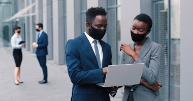 Proje yönetimi. Koruyucu maskeler takan neşeli genç iş ortakları birlikte çalışıyorlar. Yakışıklı adam dizüstü bilgisayarını tutarken çok kültürlü meslektaşı ona gülümseyerek bakıyor.