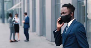 Salgın sırasında çalışmak. Koruyucu antiviral maskeli bir iş adamının portre görüntüsü akıllı telefondan konuşurken başka tarafa bakıyor ve arka planda iş arkadaşlarıyla duruyor.