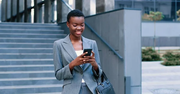 Midjan upp av multiracial dam promenader på gatan och hålla smartphone på händerna. Hon bär formella kläder medan du tittar på skärmen — Stockfoto