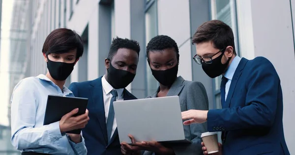 Relatório de vendas. Visão de baixo ângulo de três jovens colegas modernos usando máscaras protetoras conduzindo uma apresentação de negócios enquanto ficam na rua e olhando para o laptop juntos — Fotografia de Stock