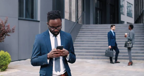 Jag hör alltid av mig. Kvalificerad ung optimistisk multiracial man bär skyddsmask läser e-post med hjälp av smartphone. Han uttrycker glädje när han står med gadget på gatan — Stockfoto