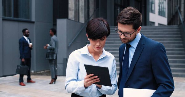 Работаем вместе. Рядом с офисным центром стоят веселые стильные молодые коллеги в формальной одежде. Привлекательная женщина держит планшет, в то время как белый мужчина жестикулирует с улыбкой