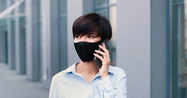 Elegante ervaren volwassen blanke zakenvrouw in formele kleding met een beschermend masker loopt op straat en kijkt bedachtzaam terwijl ze aan het chatten is op haar smartphone tijdens haar pauze — Stockfoto