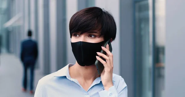 Elegante erwachsene kaukasische Geschäftsfrau in formeller Kleidung mit Schutzmaske geht auf die Straße und schaut nachdenklich auf ihr Smartphone, während sie in der Pause plaudert — Stockfoto