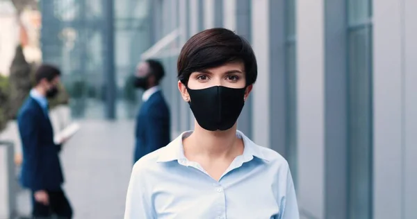 Trabajo durante la epidemia. Retrato de la mujer de negocios con máscara antiviral protectora mirando a la cámara mientras está de pie con sus colegas en el fondo — Foto de Stock