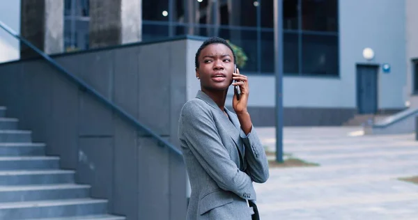 Attractive, good-wearing adult woman in suit standing nearby business center. She looking aside, smiling and holding telephone in hand while having pleasure conversation