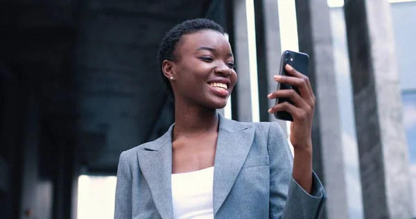 Rozhovor s potěšením. Portrét šťastné mladé kvalifikované multiraciální žena v obleku stojí na ulici a držení mobilního telefonu, zatímco s video chat — Stock fotografie