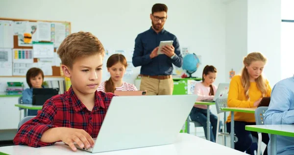 Close-up portret van schattige blanke tiener jongen leerling typen op laptop browsen online studeren op de computer wetenschap klasse, basisschool les. Man leraar helpen kinderen met behulp van tablet op de achtergrond — Stockfoto
