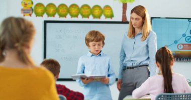 Sınıfın ve güzel bayan öğretmenin önünde dikilirken kitap okuyarak soruları cevaplayan küçük beyaz bir öğrenci. Ev ödevi. Eğitim, ilköğretim