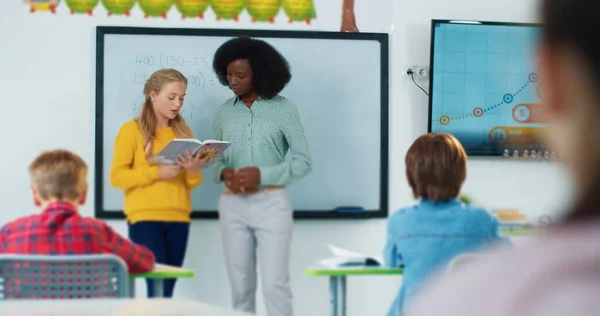 Tiener schattig blank junior student meisje staan in het midden van de klas en het beantwoorden van vraag tijdens het lezen uit leerboek in het bijzijn van klasgenoten en mooie jonge Afro-Amerikaanse vrouwelijke leraar — Stockfoto