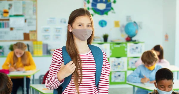얼굴 마스크를 쓴 행복 한 백인 여학생의 모습 이 코로나 바이러스가 유행하고 있는 지역 초등 학교 학생들 이 코로나 바이러스에 감염되어 교실에 서서 웃고 있다. — 스톡 사진