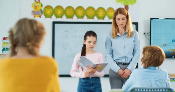 Pequeña linda colegiala caucásica que responde a la pregunta leyendo un libro de texto mientras está de pie en medio del aula frente a compañeros de clase y una bonita maestra. Estudiar, educación primaria — Foto de Stock