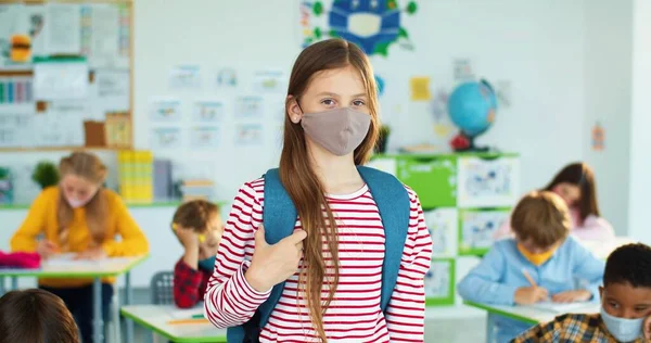 Retrato de una niña caucásica feliz usando mascarilla en el aula y sonriendo a la cámara en el interior Estudiando después de la pandemia de coronavirus alumnos de primaria multiétnica aprendiendo durante covid —  Fotos de Stock