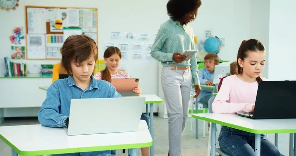 Barn lär sig i grundskolan. Porträtt av kaukasiska skolflicka och skolpojke skriva på bärbar dator surfa internet på dator vetenskap klass afroamerikanska kvinnliga lärare knacka på surfplatta bakom — Stockfoto