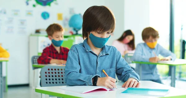 백인 꼬마밝은 학생 이 책상에 앉아서 수업중에 글을 쓰고 손을 들고 질문에 답하고 싶어 합니다.. — 스톡 사진