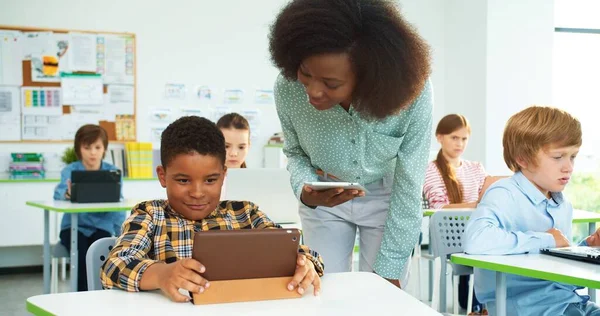 Retrato de cerca de una joven y hermosa profesora afroamericana ayudando a un colegial en la lección, educación primaria, clase de ciencias de la computación. Estudiante junior tocando en la tableta en la lección — Foto de Stock