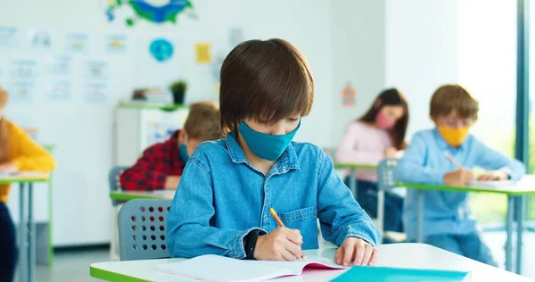 Portrét malého bílého školáka v masce, sedícího za stolem ve třídě a zapisujícího si na lekci a zvedajícího ruku, chce odpovědět na otázku. — Stock fotografie