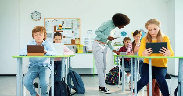 Afro-americano jovem bonita professora com gadget ensinar alunos na escola. Estudantes juniores caucasianos meninos e meninas aprendendo na aula de informática da escola usando tablets e laptops — Fotografia de Stock