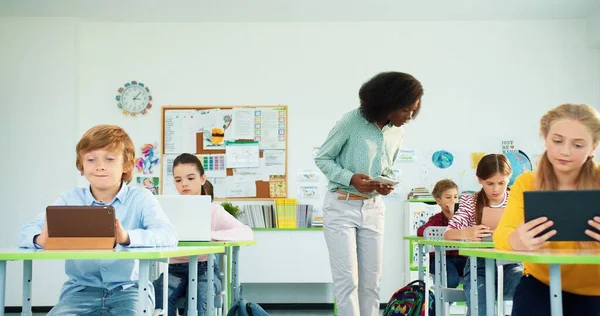 非洲裔美国年轻漂亮的女教师，在学校里带着教学用具。使用平板电脑和膝上型计算机装置在学校学习的高加索小学男女学生 — 图库照片