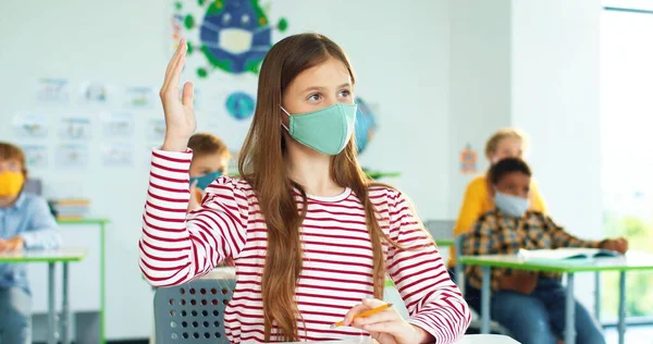 학교 책상에서 마스크를 쓰고 앉아 있는 백인 여학생의 사진을 닫아. 혼합 인종의 학생들 이 교실에서 손을 들고 있습니다. 코로나 바이러스 유행병, 교육, 창의적 사고 — 스톡 사진