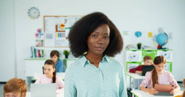 Close-up portret van mooie jonge Afro-Amerikaanse vrouwelijke leraar die in de klas staat en naar de camera kijkt en glimlacht. Kinderen die laptop en tablet op de achtergrond gebruiken. Vrouwelijke leraar op school op het werk — Stockfoto