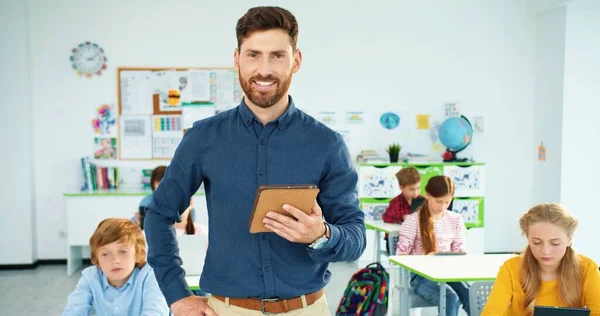 Retrato de un guapo joven y alegre profesor caucásico de pie en el aula escribiendo en la tableta, mirando a la cámara y sonriendo. Niños aprendiendo en el portátil y dispositivos en segundo plano. Educación escolar —  Fotos de Stock