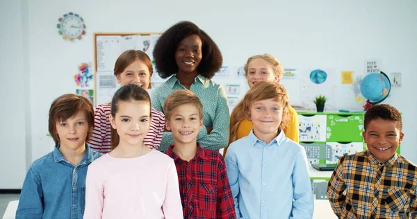 Retrato de alumnos multiétnicos alegres y diversos niños y niñas en el aula mirando a la cámara con sonrisa en las caras de pie con la joven maestra afroamericana. Niños felices con tutor en la escuela —  Fotos de Stock