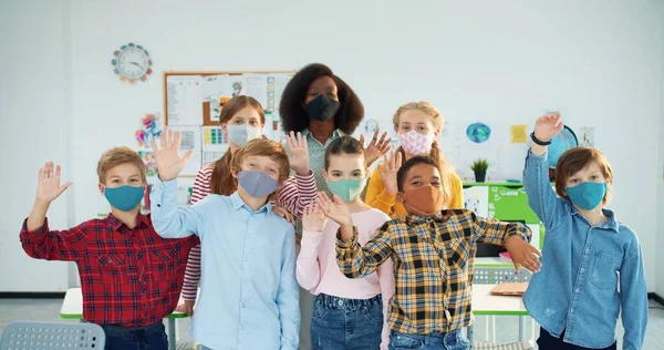 Portret mieszanej rasy radosnych dzieci w maskach stojących w klasie żeńskiej w szkole z młodą, ładną afroamerykańską nauczycielką patrzącą w kamerę, uśmiechniętą i machającą dłońmi podczas pandemii — Zdjęcie stockowe