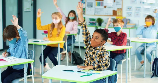Różnorodne rasy mieszane dzieci siedzące w klasie ucząc się i podnosząc ręce gotowe do odpowiedzi na zagadnienie Szczęśliwi mali inteligentni uczniowie szkoły podstawowej uczący się w szkole podczas edukacji covid — Zdjęcie stockowe