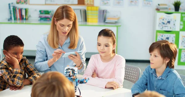 Biała młoda, piękna blondynka nauczyciel kontroluje robota elektronicznym gadżetem za pomocą smartfona. Dzieci z nauczycielem na lekcji budowy robotów. Zajęcia z robotyki i wynalazków — Zdjęcie stockowe