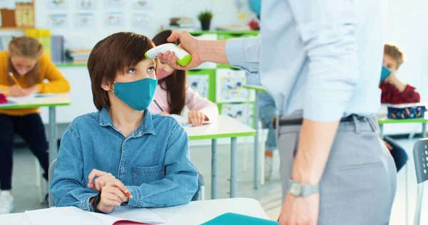 Las maestras dan la mano en el aula midiendo la temperatura del estudiante de primaria caucásica con máscara sentada en el aula en la lección usando termómetro infrarrojo durante el covidio-19. Primer plano retrato —  Fotos de Stock