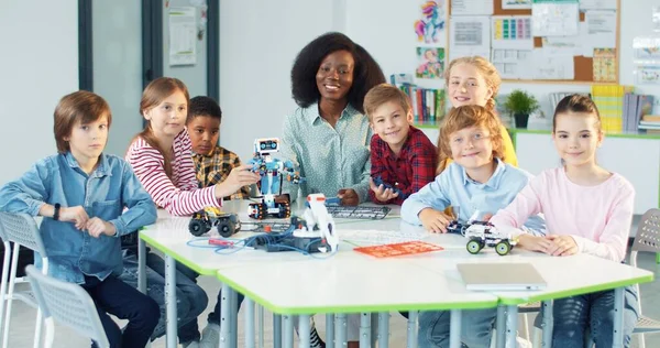 Felices niños multiétnicos sentados en el aula con la joven negra profesora guapa mirando a la cámara y sonriendo. Clase de ciencias de la tecnología, estudiar en la escuela primaria, educación moderna — Foto de Stock