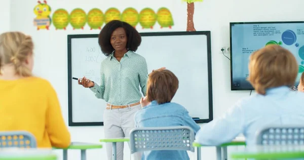 Szczęśliwa Afroamerykanka piękna młoda nauczycielka stojąca obok tablicy wyjaśniającej lekcję dzieciom w klasie i zadająca pytania, a dzieci podnoszą ręce. Kształcenie podstawowe — Zdjęcie stockowe