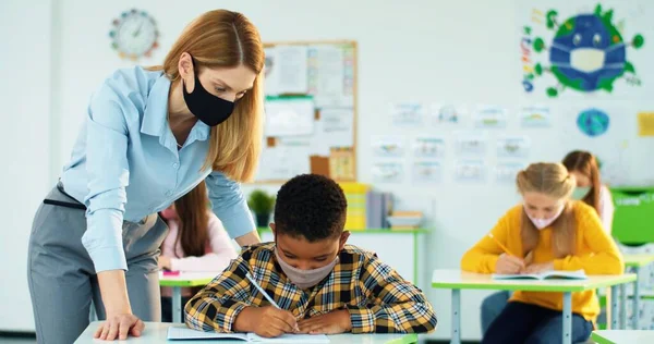 Retrato de una joven y hermosa profesora caucásica ayudando a un pequeño estudiante afroamericano a escribir una prueba en copybook en clase. Mujer enseñando brillante colegial explicando lección Post-covidio —  Fotos de Stock