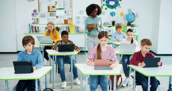 Afroamerykanka ładna nauczycielka w szkole chodzi w klasie używając tabletu na lekcjach. Dzieci rasy mieszanej, młodzież ucząca się przy biurkach, pracująca nad urządzeniami, studiująca technikę — Zdjęcie stockowe