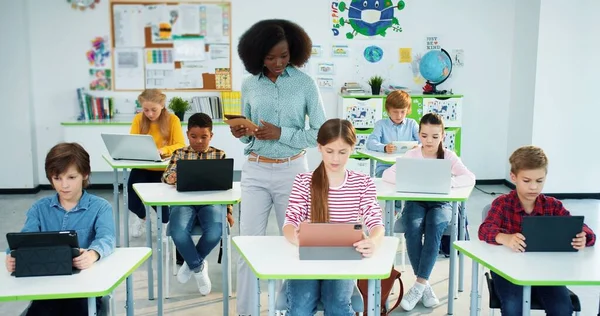 Multi-etnické rozmanité junior studenti studují ve škole pomocí notebooku a tablet, technologie, počítačové vědy třídy. Africký americký mladý fena učitel ve škole výuka světlé třídy žáků — Stock fotografie