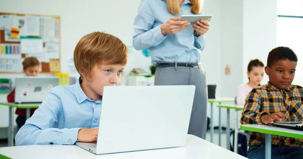 Close-up van een blanke kleine basisschoolstudent die achter een bureau zit in de klas, typend op laptop tijdens de les, een vrouwelijke leraar die achter een tablet loopt. Leren van gemengde rassen in computerklassen — Stockfoto