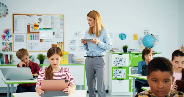 Basisschool informatica klas. Slimme multi-etnische junior studenten meisjes en jongens gebruiken laptop. Vrouwelijke leraar legt les uit met behulp van tablet. Internettechnologie. Modern onderwijsconcept — Stockfoto