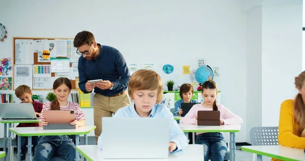 Clase de informática en la escuela primaria. estudiantes junior niñas y niños utilizan el ordenador portátil en la lección. Profesor masculino explicando y ayudando a la colegiala en clase usando tableta Tecnología de Internet Educación moderna —  Fotos de Stock