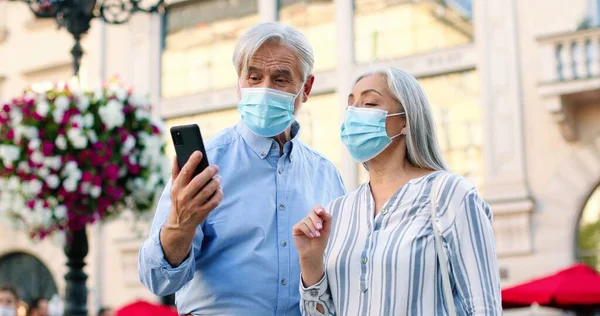 Hej. Låg vinkel syn på senior pension par bär skyddsmasker tittar på smartphone skärmen och hålla videosamtal med någon när du går under pandemi — Stockfoto