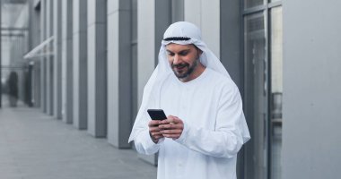Kandora giyen Arap işadamının portre görüntüsünü belden yukarı kaldırın. Cep telefonuna bakın ve başarılı iş anlaşmasından mutlu olurken sevinin.