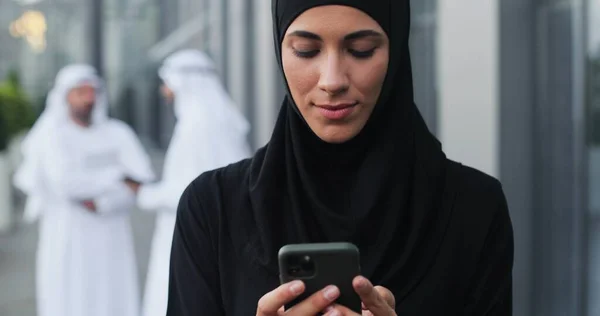 Використання смартфона. Очікувати портрет арабської бізнес-леді в шарфі голови, пишучи серйозне повідомлення комусь, тримаючи її мобільний телефон на вулиці — стокове фото